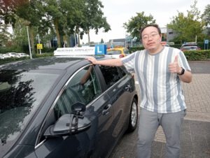 A jubilant man standing next to his learner car, celebrating passing his driving test in Eindhoven with a big smile.